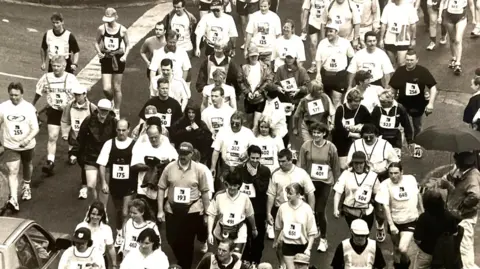 Manx National Heritage A camera is high up above the heads of those walkers who have started the parish walk. Everyone is wearing numbers on their tops. There are around 40 people in the photograph. 