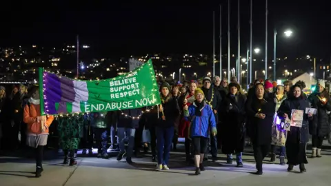 Penzance Council The photo shows people marching down a promenade in Penzance at night. There is a green banner that says "end abuse now".
