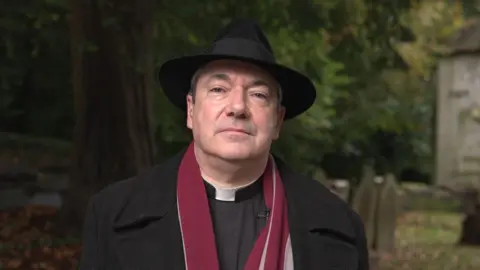 BBC A man wearing a trilby, long black coat, and burgundy scarf stands beneath a tree, in front of several aged headstones.