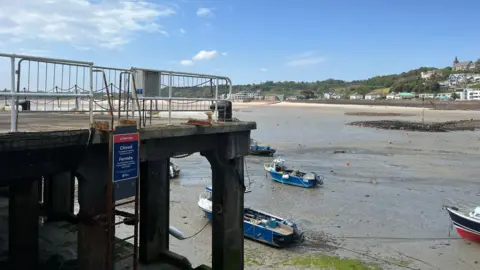 BBC The image shows a small concrete pier with metal railings. It is low tide. There is a ladder running down the side of the pier, and a sign indicating the pier is closed. Several blue and white fishing boats rest on the exposed muddy harbour bottom below. Across the water, there is a sandy shoreline and a hillside dotted with houses stretch under a bright blue sky with scattered clouds.