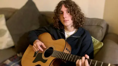 BBC Alex Spencer with long curly hair sat holding a guitar on a sofa