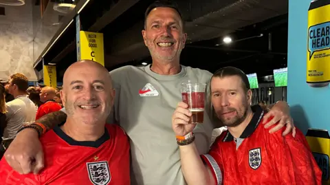 Dave Barton Three male England fans at Ashton Gate Stadium in Bristol. Two of them are sat down wearing red England football tops, while the third man is standing up between them with his arms around them. One of the men is holding a pint up.