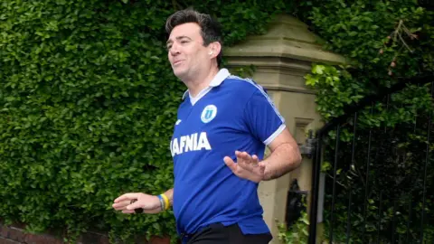PA Media Andy Burnham running near his home wearing a blue Everton shirt and black shorts. A dense wall of green foliage and hedging fills most of the background.
Behind him is a stone gatepost and a black wrought-iron gate leading into a property. A low red-brick wall runs along the edge of the pavement. 