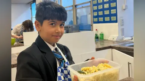 Discovery Educational Trust A young boy wears a black blazer and blue and black tie. He is holding a plastic box of pasta. He is in a school kitchen, which has mostly white-painted walls.