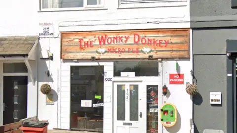 Google Frontage of the pub building- with a white door - a red bin in front and a green and yellow defibrillator on the side of the building 