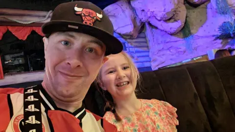 A man in a red and white football shirt and baseball cap smiles next to a younger girl in a pretty flowery dress