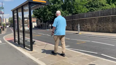 Sofía Luis-Hobbs/BBC A man holding a white mobility cane in front of him wears a a beige cap, light blue polo shirt and beige trousers as he walks in front of a bus stop. There is a cycle path to the left of the island where the bus stop is, and a road to his right.