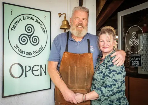 Spring Fling A man with a big beard and grey hair in a leather apron and blue T-shirt has his arm around a woman with blonde hair in a green and blue patterned top. The are standing at a shop door with a sign next to them for Triskele Artisan Jewellery