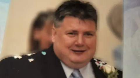 John Smyth, a well-built man with short dark hair wearing a black suit with white shirt and blue tie. he appears to have confetti scattered on his shoulders and a white flower in the lapel of of his jacket.