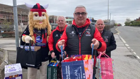 Skinn holds a bag for life crammed with easter eggs.  Two other men behind him also stand with bags as does the team mascot in a cartoon viking mask.