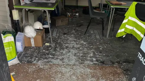 Shattered glass on the floor of a room with builders' equipment including hi-vis jackets and helmets