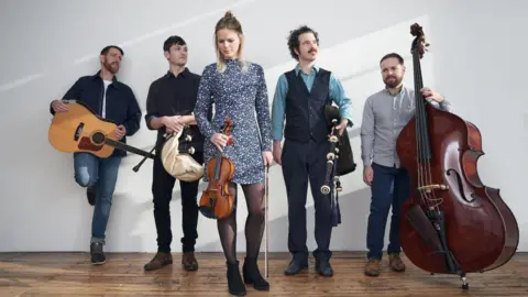 Celtic Connections Five members of the band Breabach standing on a wooden floor with their instruments. A woman in the foreground carries a fiddle while two men either side of her have bagpipes. Another man has an acoustic guitar, another a double bass. 