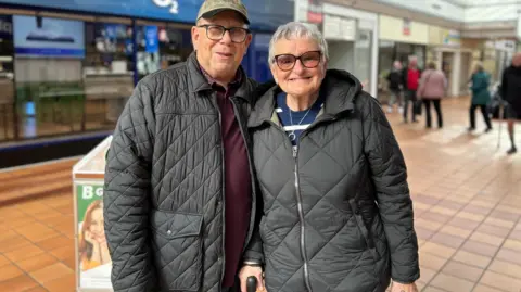 Amber Ward/BBC A married couple in their sixties pictured in a shopping centre. They are both wearing dark coats and smiling at the camera.