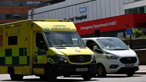 A yellow ambulance with the words London Ambulance parked next to a white van outside a hospital emergency department 