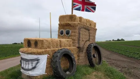 A tractor sculpture made out of straw bales. It depicts a tractor with a Union flag flying from its "cab". Two large black tyres are resting on its side. It has a face made from eyes where its windscreen would be, nostrils at the front of its bonnet and a banner painted in the shape of a smile where its bonnet grille would be. It is surrounded by fields.