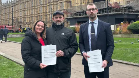 Will Stone Three people standing in front of the House of Commons, holding two copies of a bill 