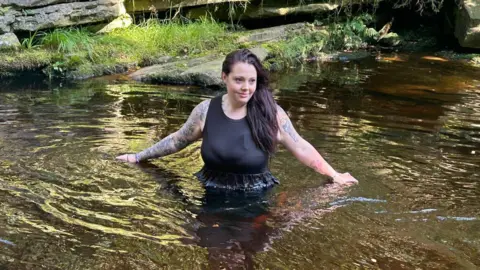 Joshua Wooldridge Bethany Wooldridge standing in a lake, with water up to her waist and her arms outstretched. She is wearing a black sleeveless dress and has tattoos on both arms. 