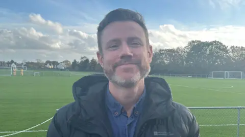 Victoria Polley/BBC Jon Waldron has short brown hair and a beard. He is wearing a coat and smiling pitchside at the Colchester United training ground.