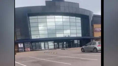 A still taken from a video, which is slightly blurry. It shows the front of the cinema with glass doors damaged, and red hazard tape across the car park. Above the main doors are glass mirrored panels.
