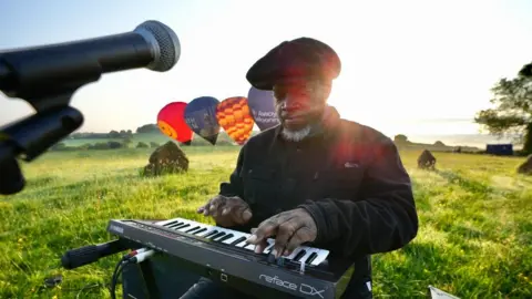 Show of Hands A man is playing a keyboard on a green field. Hot air balloons are getting ready to launch behind him. The sun is shining over his shoulder.