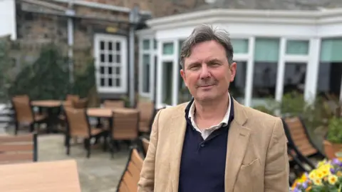 BBC The image shows Deputy Andy Sloan. He is a man with grey wavy hair and he is wearing a tweed sports jacket, a navy knitted top and a light polo shirt underneath. 