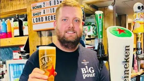 Tom stands behind a bar and holding up a tall pint glass of Heineken beer toward the camera. The glass is filled with golden lager and has a thick white head at the top. He is wearing a dark navy or black T-shirt with the words “BIG pour” and a small Heineken logo printed on the chest.