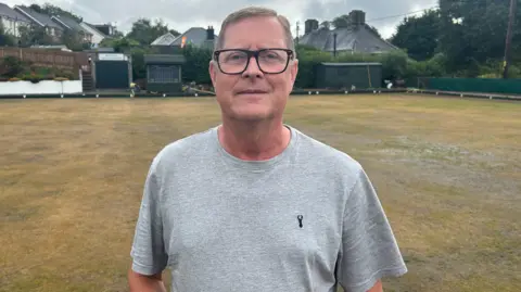 Malcolm Johnson from Ivybridge Bowls Club is stood on the bowling green and is wearing a grey t-shirt and looking straight at the camera. He is also wearing glasses.