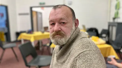 John wearing a beige jumper, with tables and chairs seen behind him