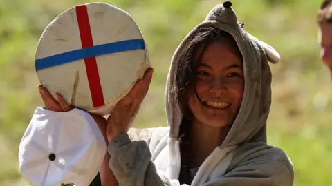Getty Images A young woman lifts a wheel of Double Gloucester cheese with blue and red bands around it in a cross. She is wearing a grubby grey hoodie with mouse ears and a snout on the hood and her hands are dirty too. She is smiling widely and has dark hair.