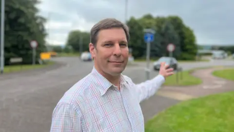 The picture shows Cllr Richard Wilkins, who's in a stripy shirt. standing next to a road in Wellington. The Chelston roundabout is behind him.