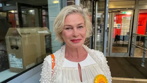 Rachel Gilmour is stood in front of the doors to a building, looking at the camera smiling. She's wearing a white top.