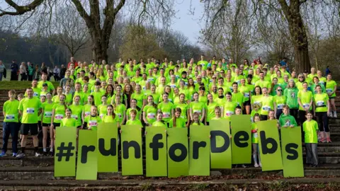 A large group of people stood on steps outside in several rows. They are wearing bright green T-shirts and are holding a sign which says 'Run for Debs'.
