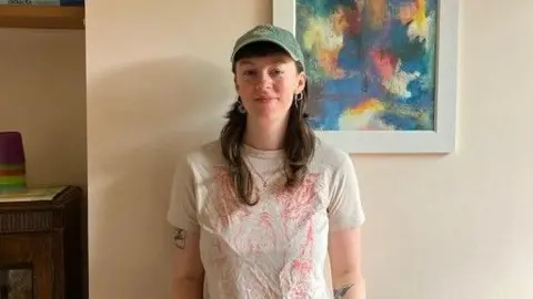 A woman standing against a cream wall. She is wearing a white top and has tattoos on her arms. She is smiling and wearing a baseball cap.