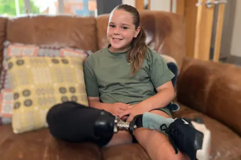 11-year-old girl smiling with her hair tied back, in a green sportswear t-shirt, with her right prosthetic leg crossed over her left leg. She is sitting on a brown leather sofa next to a yellow cushion. 