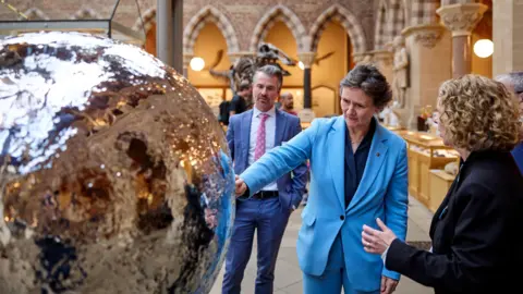 Fisher Studios and Museum of Natural History, University of Oxford Vice-chancellor Prof Irene Tracey touching the surface of a new exhibit called Mirror Moon. The museum's director, Dr Gavin Svenson, is behind her. Another woman is speaking with her.