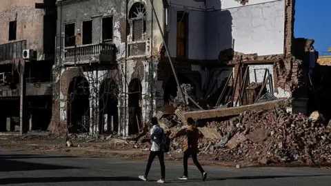 AFP via Getty Images Two men walking past a destroyed building in Khartoum. A damaged structure is still standing on the left but on the right, the building is reduced to rubble