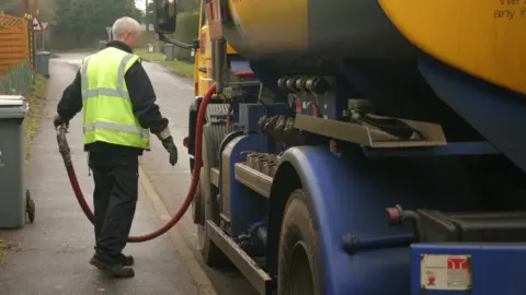 A delivery driver pulls the hose out of a small oil tanker lorry as it is parked by the side of a residential street.