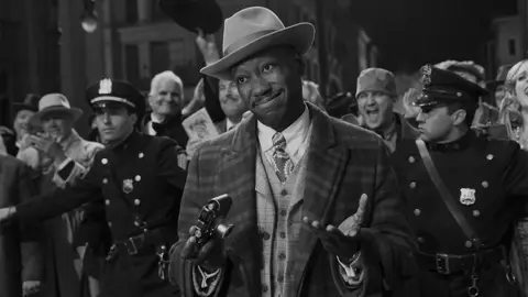 Aaron Epstein/Prime A black and white image of the character of Robbie Robertson, wearing a hat and holding both hands out while in a crowd of people, who are behind him, and being held back by police. 