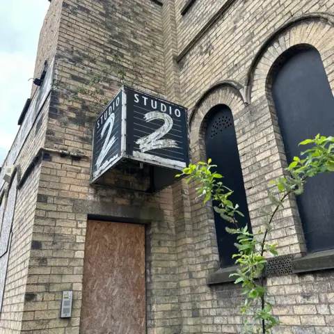 BBC/James Piekos A sign in the shape of a black cube is attached to a yellow-brick wall above a boarded-up doorway. White text on two sides reads "Studio 2". To the right, branches with green leaves grow in front of two tall, arched windows, which are covered in black boards.