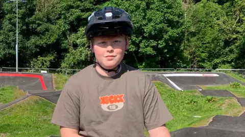 A young man in a brown T-shirt with a black helmet on stands in front of a bicycle pump track