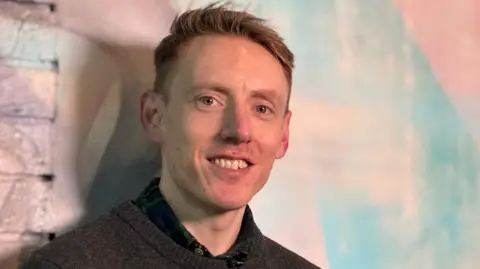 A clean-cut man in his thirties is looking straight at the camera and smiling. We can just see a dark sweater and a black shirt. He's in front of a wall painted a colourful mix of lights pinks and blues 