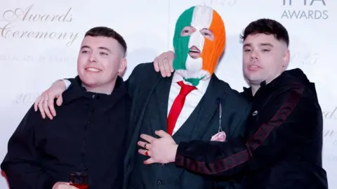 Members of Kneecap pose on the red carpet at the Irish Film and Television Academy (IFTA) Awards. Two band members are dressed in black tracksuits and have short dark hair. One member is dressed in a black suit with a white shirt and red tie and a green white and orange balaclava.