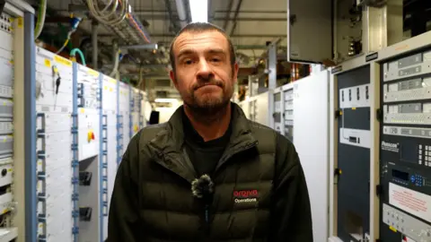 A man wearing a dark Arqiva Operations jacket stands inside a busy technical room filled with tall racks of broadcasting equipment. Rows of metal cabinets and electronic panels line both sides of the corridor, covered in buttons, switches and indicator lights.