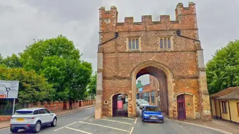 South Gate in King's Lynn, with cars driving through the area. Trees are lining the road with a road sign on the left hand side