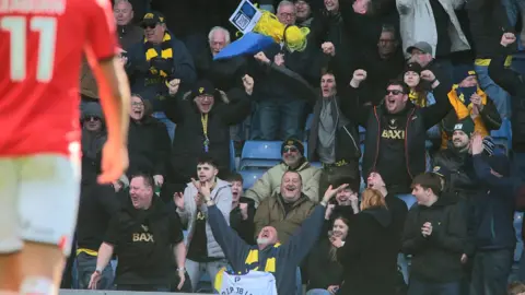 Gary Hudson throws his yellow wig and blue beard in the air in celebration, as the Oxford fans around him also celebrate.