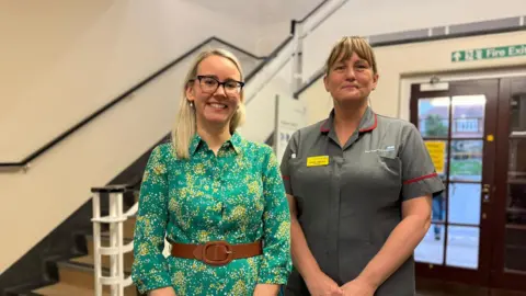 A woman wearing black framed glasses and a green dress with a yellow pattern stands next to her colleague who is wearing a grey NHS branded shirt. 