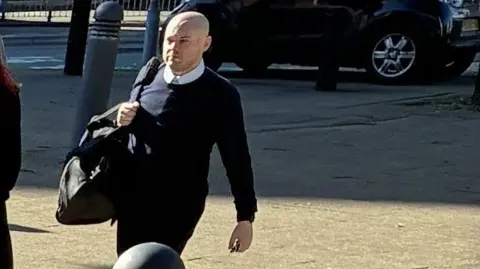 Adrian Hughes arriving at Preston Crown Court. He is wearing a dark blue jumper, dark trousers, and carrying a black bag. He has a bald head. 
