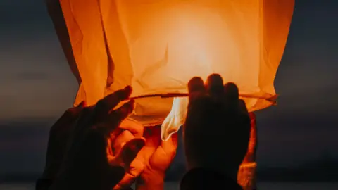 Getty Images Hands releasing a lit sky lantern. There is a flame at the bottom and the lantern is glowing orange.