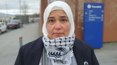 Majida Al Askri looks forward. She has a white head scarf on and a black and white palestinian keffiyeh on her neck. She is wearing a black blazer with a shamrock badge on it.