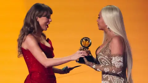 Taylor Swift, dressed in red, hands Beyonce, in gold, a trophy 